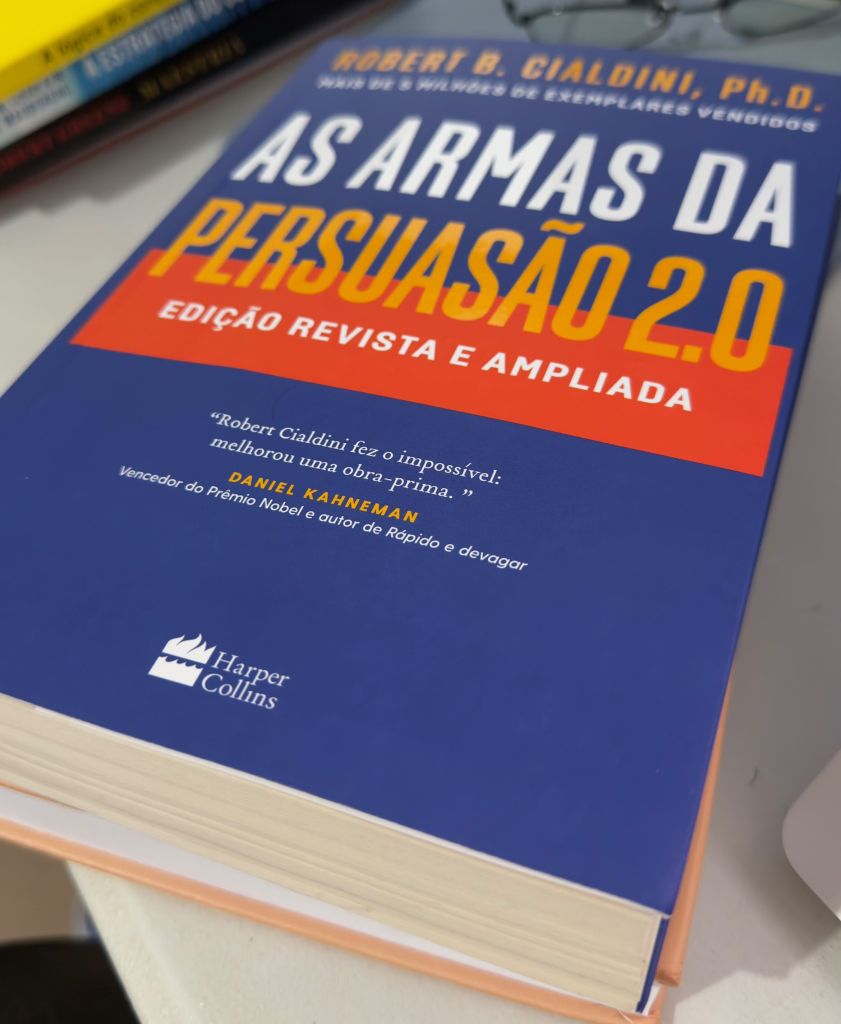 Exemplar de As armas da persuasão 2.0, de Robert Cialdini, sobre uma pilha de livros