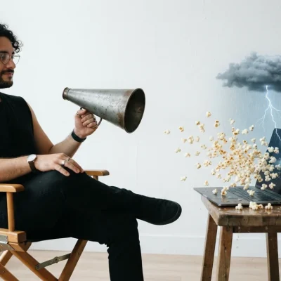 Homem em cadeira de diretor dá ordens com um megafone a um notebook, de onde saem chuva, um raio e pipocas estourando